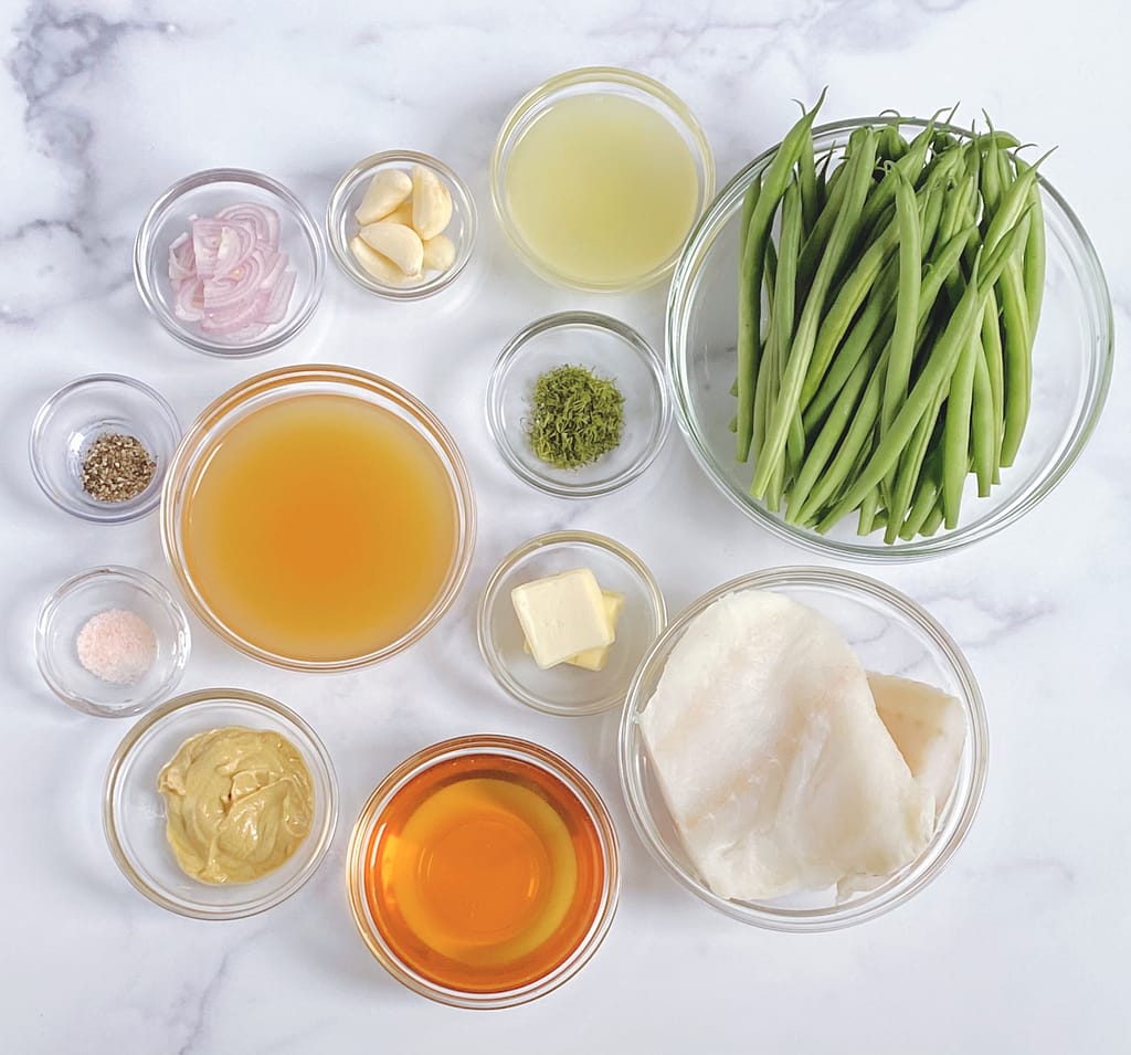An overhead view of all the ingredients for Cod with Green Beans and Shallot Dill Lemon Butter Sauce, in individual bowls: cod, green beans, fresh dill, dijon mustard, white wine, garlic cloves, sliced shallors, lemons, chicken stock and salt & pepper