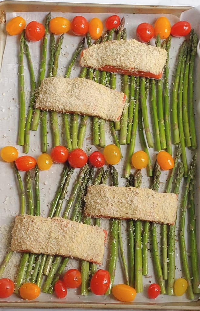 Overhead view of raw salmon filets with sauce and panko on top, cherry tomatoes and asparagus spread out on a sheet pan