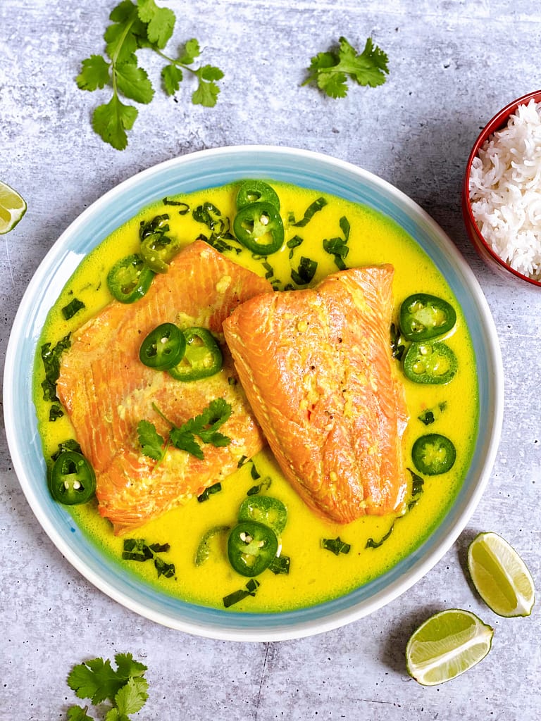 Overhead view of plated Spicy Coconut Lime Salmon with a bowl of cooked jasmine rice in the upper right hand corner. Sliced limes and basil are on the table.