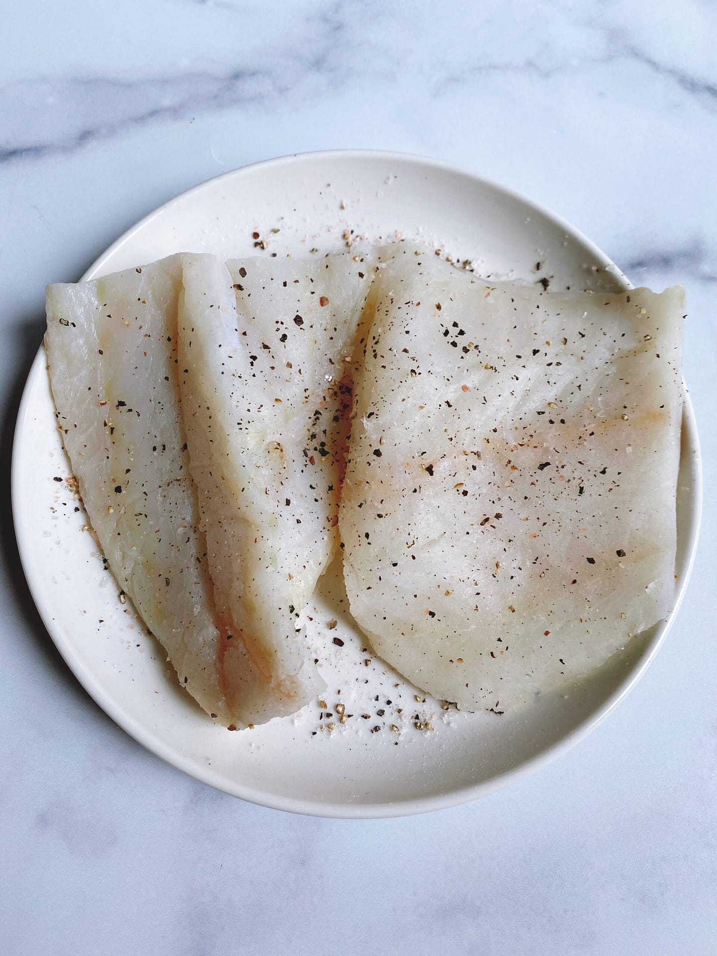 A plate of two raw cod filets seasoned with salt and pepper.