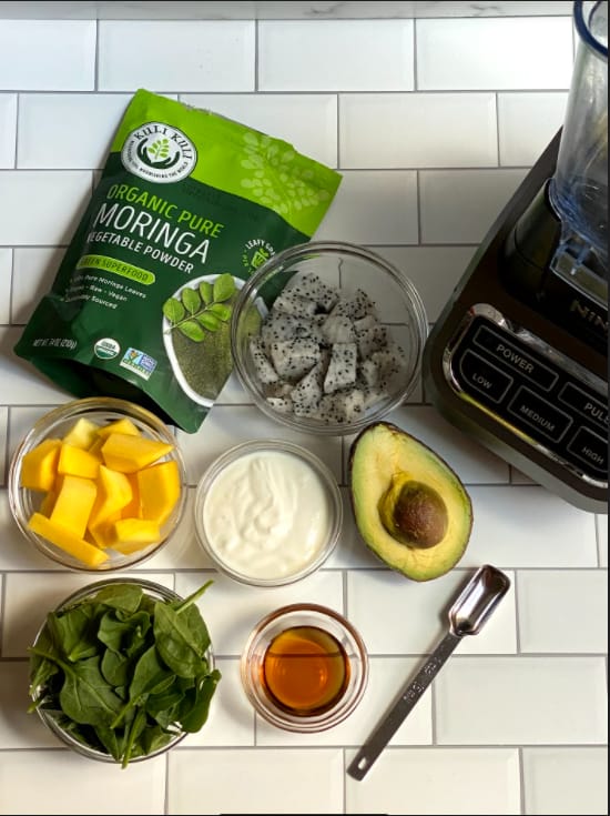 Overhead view of all the ingredients for the Mango Moringa Superfood Smoothie: a bowl of mangos, a bowl of spinach, a bowl of Greek yogurt, a bowl of dragon fruit, half an avocado, a bowl of honey