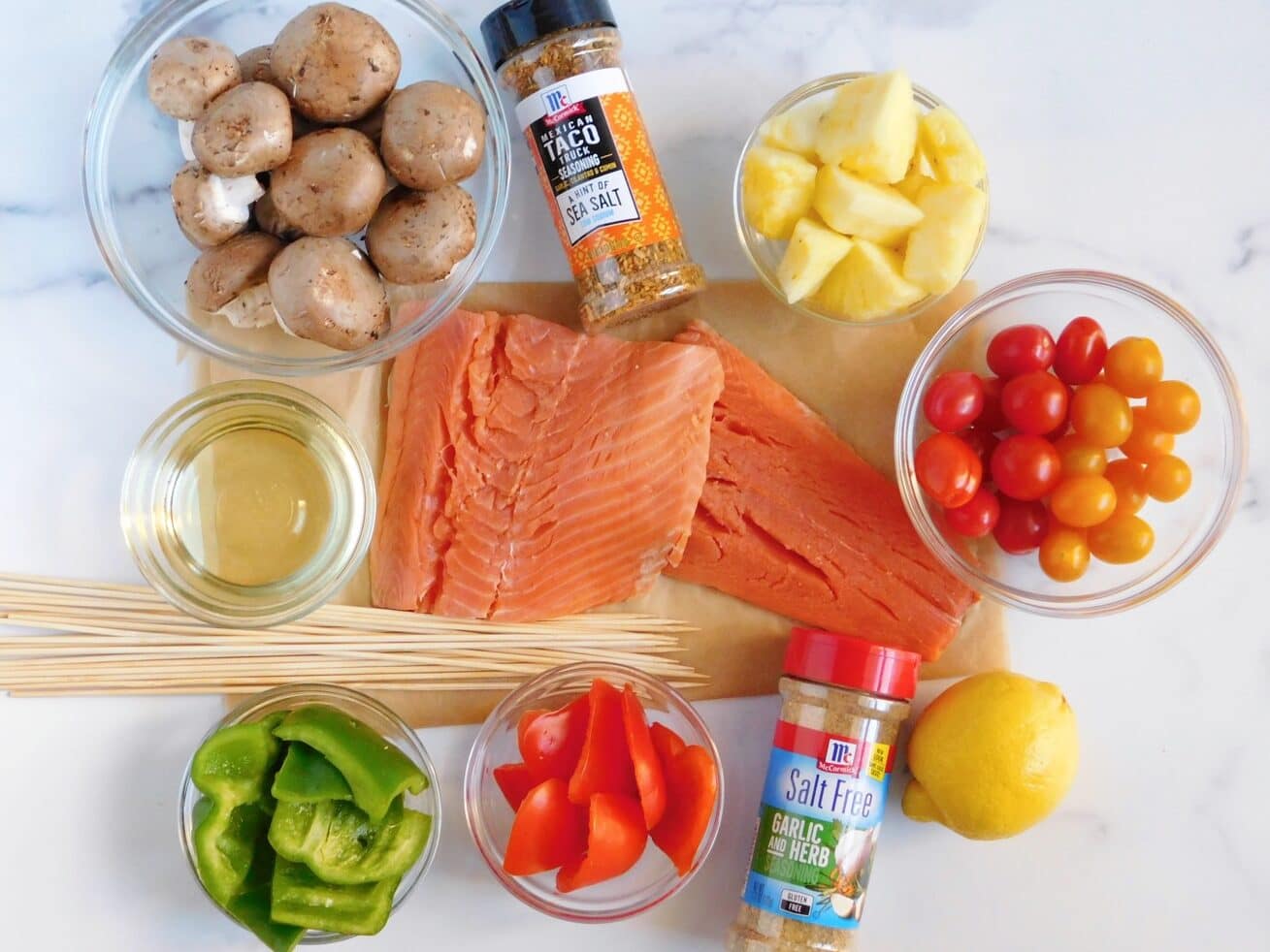 Overhead view of ingredients for Grilled Salmon Kabobs including: a bowl of cremini mushrooms, McCormick Mexican Taco Food Truck Seasoning, a bowl of sliced pineapples, a bowl of tri-colored tomatoes, one whole lemon, McCormick Salt Free Garlic & Herb Seasoning, sliced red and green peppers in separate bowls, an assortment of bamboo skewers, a bowl of vegetable oil and two salmon filets in the center.